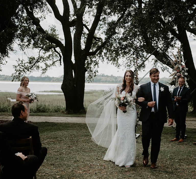 Bride in floral lace wedding dress and groom in black suit and blue tie walk back down the aisle as a married couple 