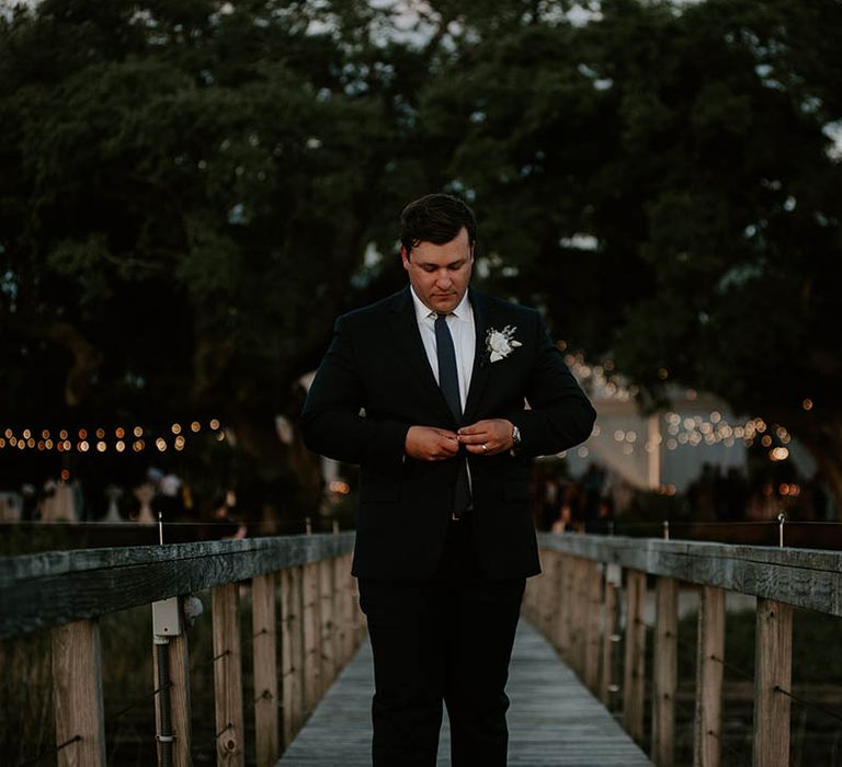 Groom in black suit with white shirt and blue tie wearing white rose buttonhole for wedding 