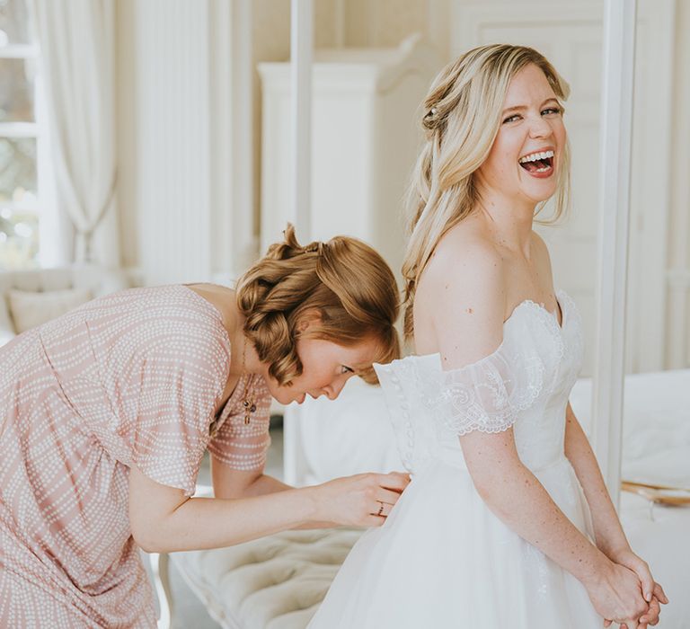 Bridesmaid buttons up the bride in her lace off the shoulder wedding dress with long blonde hair 