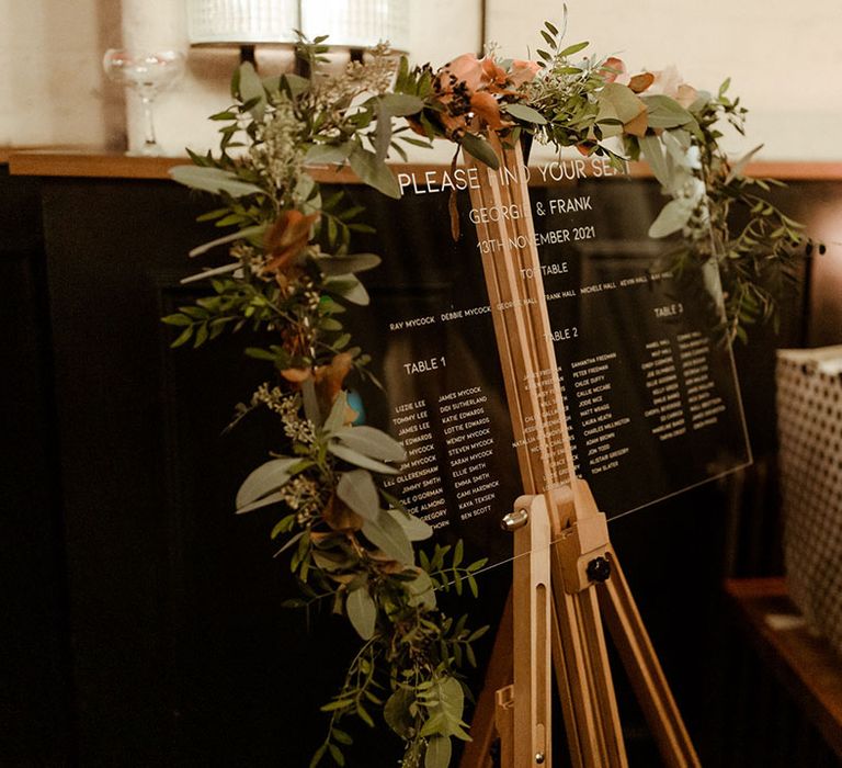 Wooden easel complete with glass tableplan surrounded by florals and green foliage 