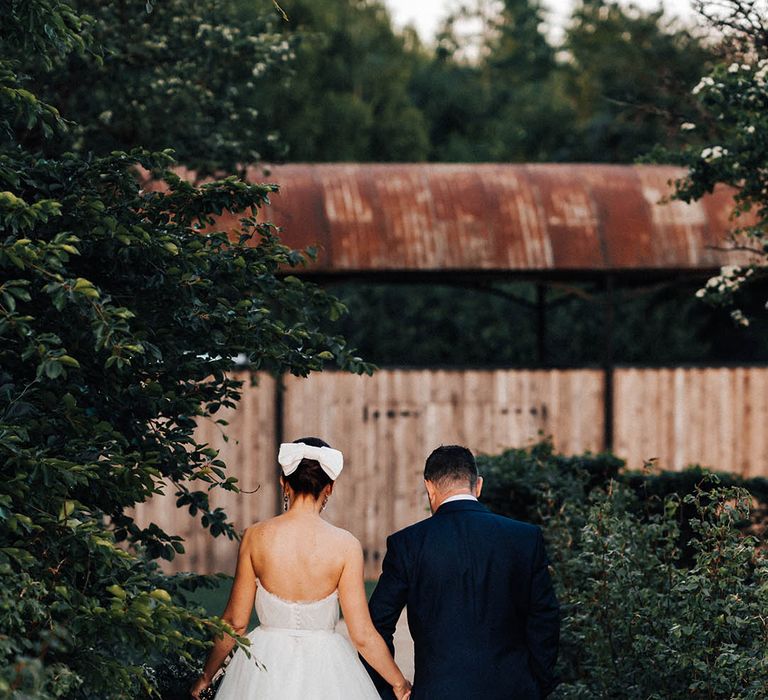 Bride in strapless backless wedding dress with ruffles and tulle princess skirt holding hands with groom in blue suit 