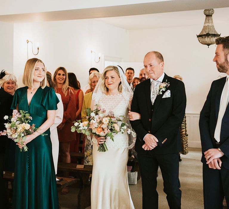 Bride walks down the aisle with her father whilst carrying pastel coloured floral bouquet 