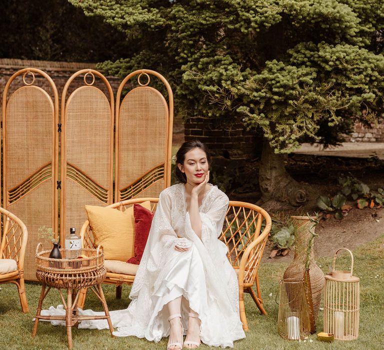 East Asian bride in a satin slip wedding dress with lace kimono style overlay sitting on a bamboo chair surrounded by a bamboo screen and table 