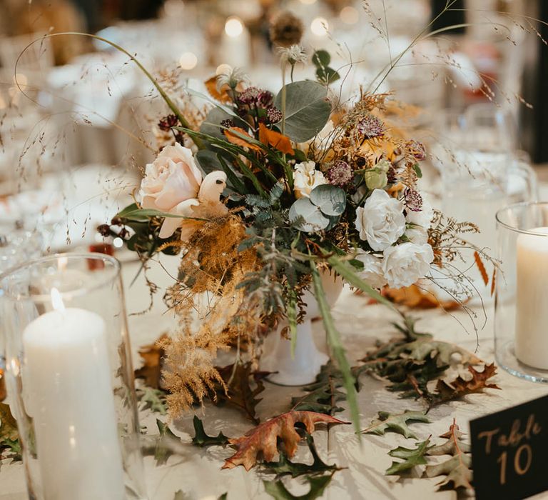 Wedding table centrepiece with autumnal leaves, white roses and dried grasses with white candles 