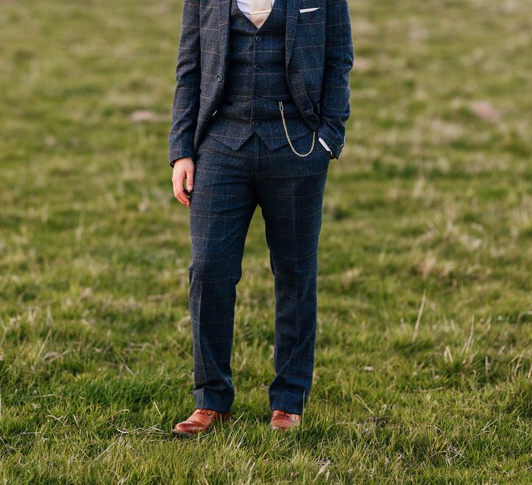 Groom in three piece blue checkered suit with pale tie with gold tie clip with brown shoes 