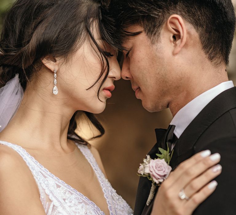 Bride wears diamond drop earrings and places her forehead against her groom wearing black tie and floral buttonhole 