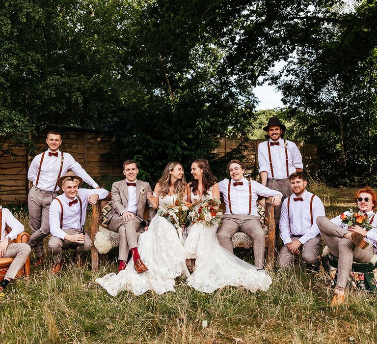 Bride sit outside together with all their groomsmen in orange bow ties and braces in grey trousers and suit jackets 