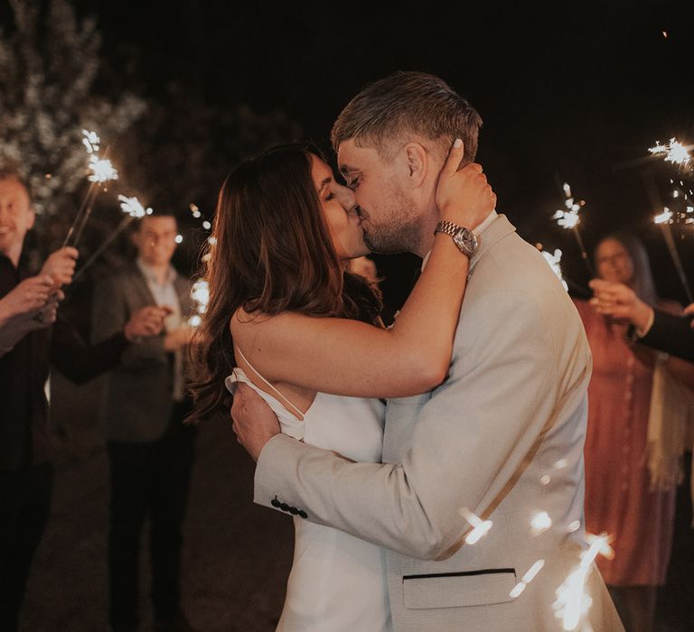 Bride and groom share a kiss as guest wave sparklers in celebration 