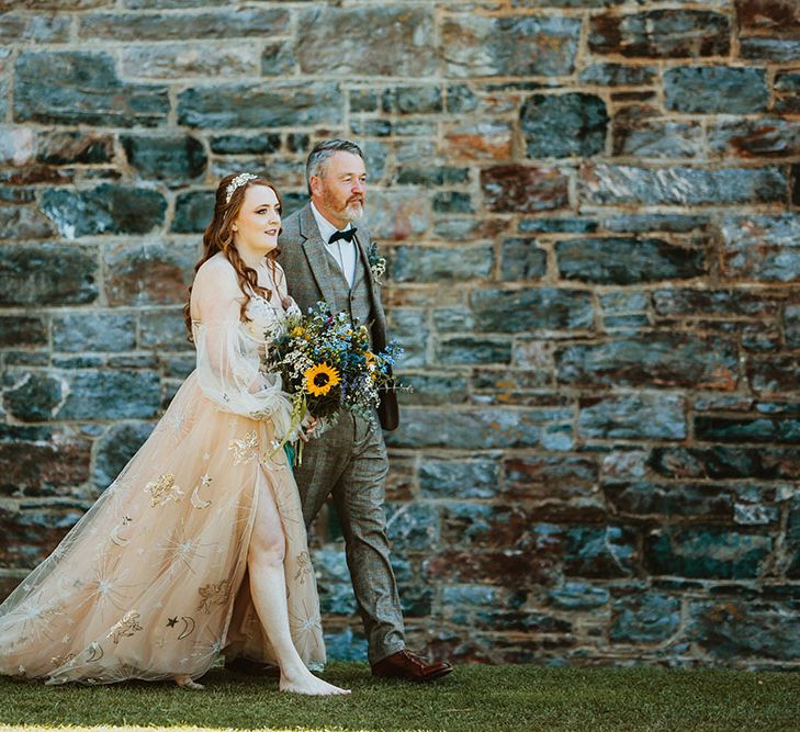 Bride wearing pink starry wedding dress walks with her father who wears three piece tartan suit with black bow tie and white shirt