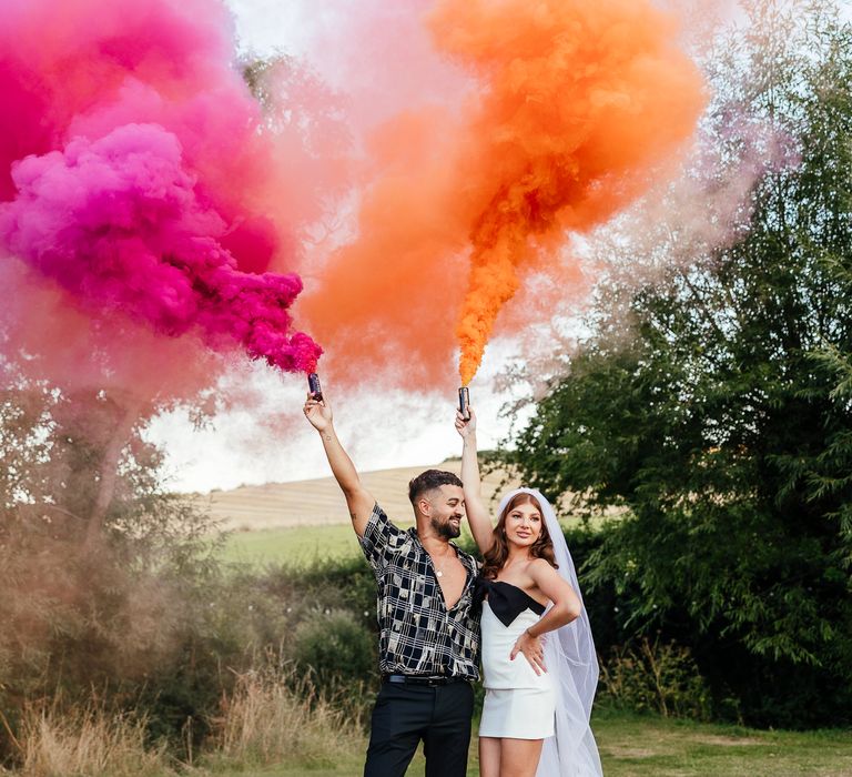 Bride & groom hold orange and pink smoke bombs outdoors for couples portraits 