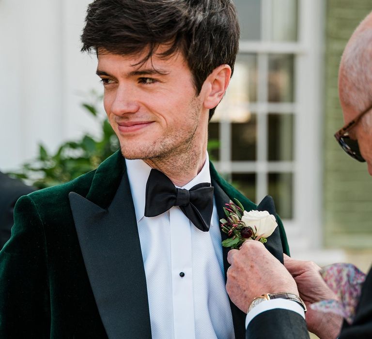 Groom wears black tie and green velvet suit jacket with white rose buttonhole tied with white ribbon 