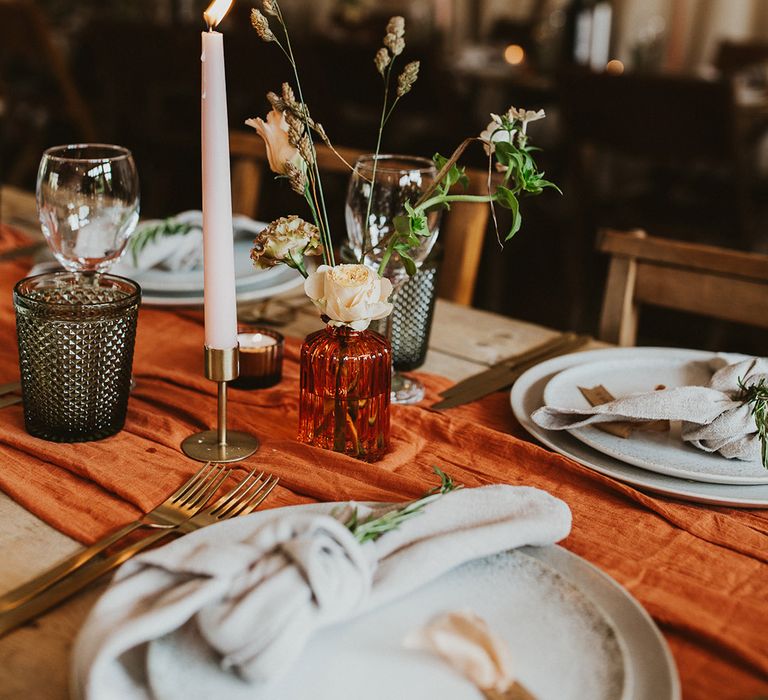 Flower stems in orange vase with candles, an orange table runner, and white plating 