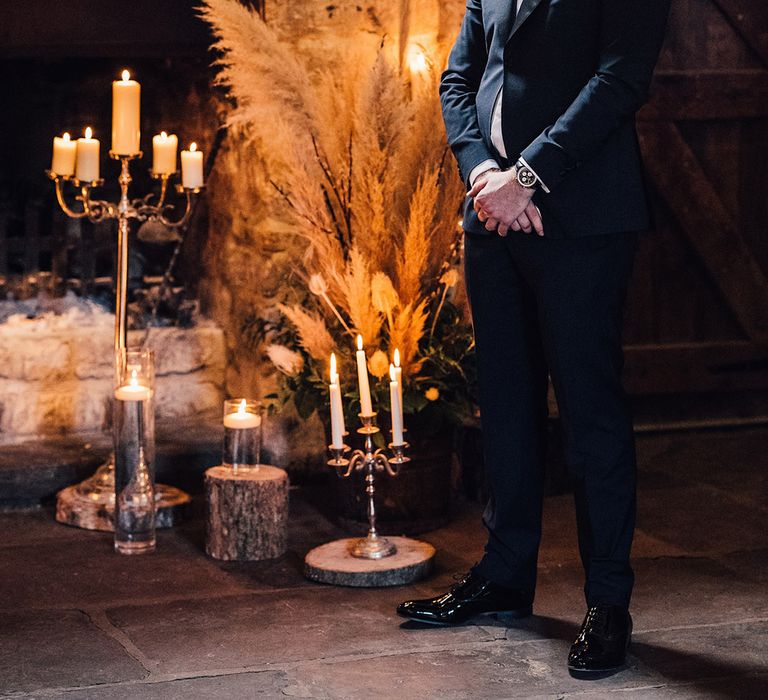 Groom in black tie turns to look at the bride as she enters with pampas grass decor and tall gold candelabras