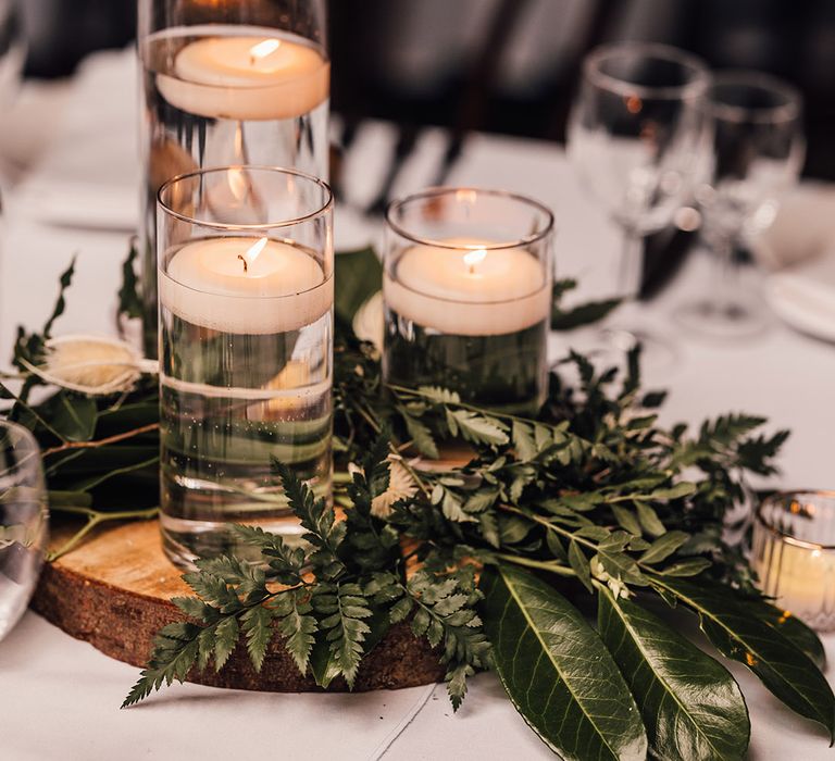 Hurricane vases full of water and a floating tea light with green foliage on a tree trunk slice for the table centrepieces 