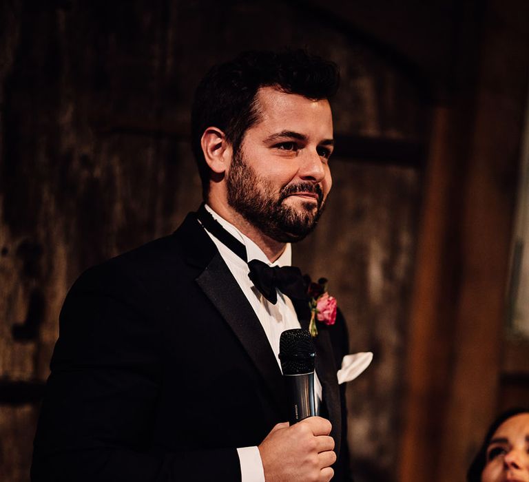 The groom in traditional black tie stands up with a microphone as he reads out his speech 