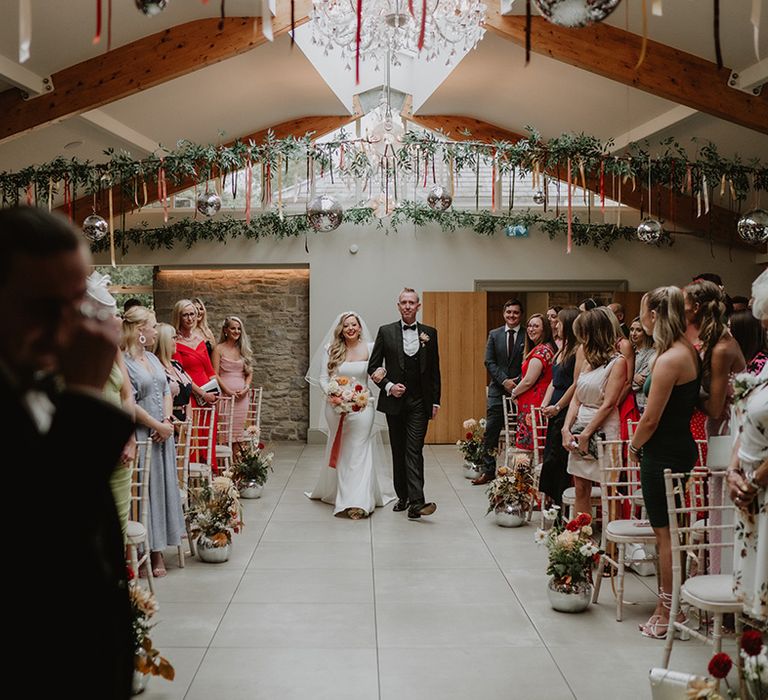 Bride in square neck satin wedding dress with two tier veil walking up the alter with short colourful streamers, disco balls, a backdrop banner and foliage decorations hanging 