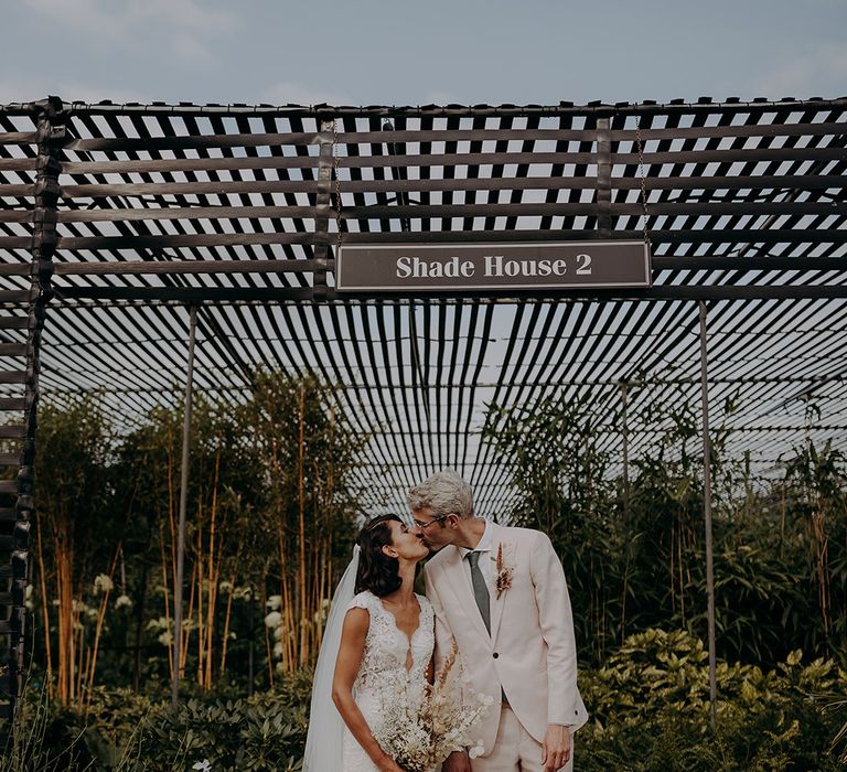 Bride and Groom kiss in front of greenhouse wearing a lace and tulle fishtail dress and a light pink suit