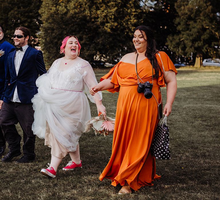 Bride in corset top wedding dress with leaf detailed lace overlay, pink stitching around the waist and red gemstone necklace and red vans walking with bridesmaid in orange off the shoulder bridesmaid dress and groom in midnight blue blazer with galaxy patterned bowtie and autumnal boutonniere as well as best man in midnight blazer and white shirt with sunglasses