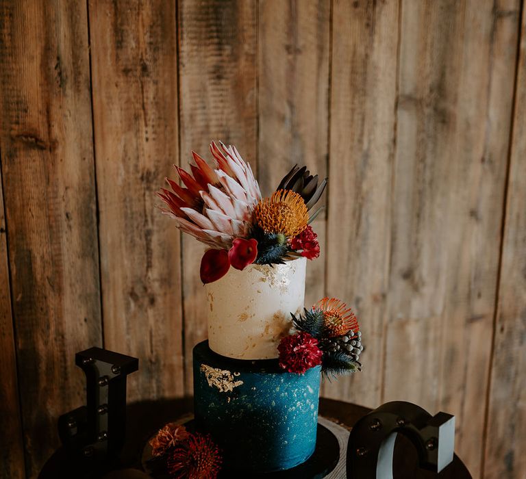 Two tier deep blue layer and white layer with gold flakes with king proteas and orange flowers 