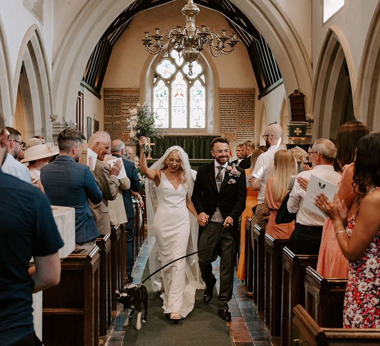 The bride and groom walk back down the aisle as a married couple with their pet dog, Brains 