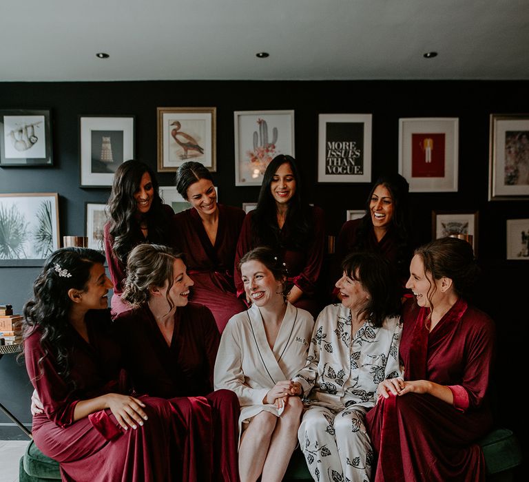 Bridesmaids in dark red satin robes, bride in cream robe with black stitching and mother of the bride in floral patterned silk pyjamas getting ready before wedding