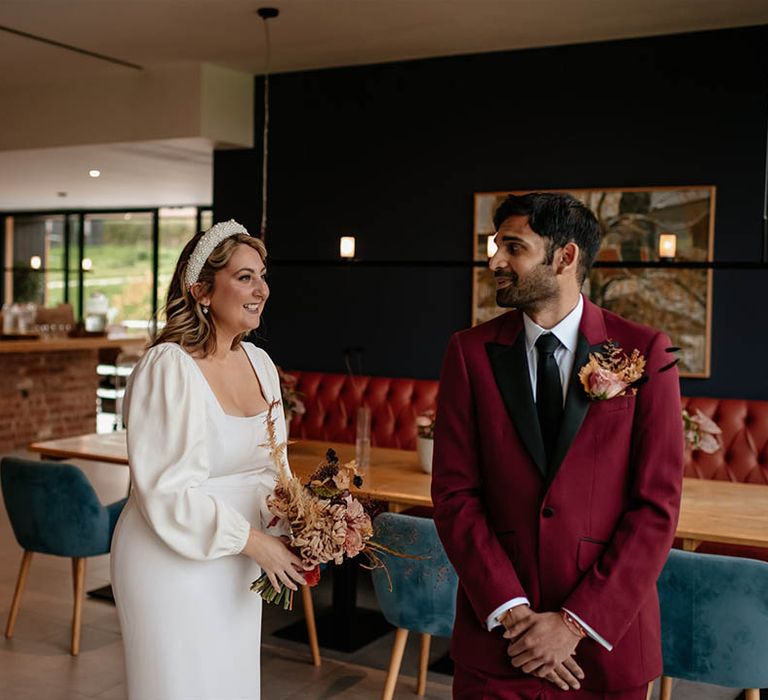 Bride wearing a long sleeve wedding dress with square neckline and pearl headband walks in for a first look with the groom in a deep red suit jacket