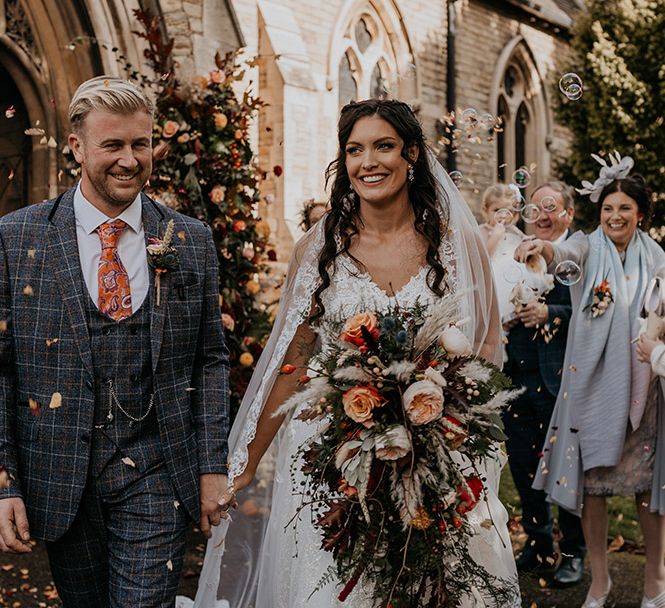 Groom in three piece grey checkered suit walking with the bride in a lace wedding dress as they have their confetti exit with the bride holding a boho cascade wedding bouquet