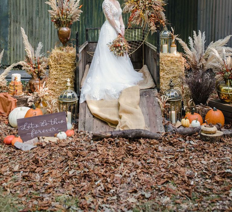 Bride in long sleeved v neck soft ivory dress with lace floral detailing on the bodice and sheer sleeves standing on the back of a wooden trailer at harvest wedding with giant gold crescent moon ceremony backdrop and eco-friendly sustainable wedding decor, large dried florals, pumpkins and calligraphy signage