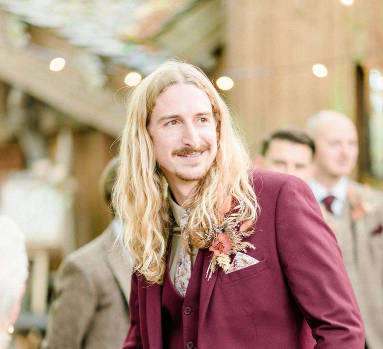 Groom wears maroon suit finished with floral pocket square and matching tie and dried floral buttonhole