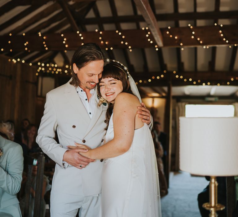 Bride in silk v-neck sleeveless ASOS wedding dress and gold halo headband with sparkly stars smiling and hugging groom in neutral coloured double-breasted ASOS suit