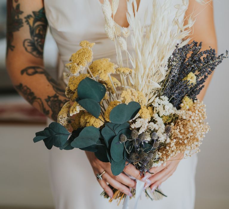 Bride holding dried flower wedding bouquet with white bunny tails, green foliage, yellow dried gypsophila, lavender and baby’s-breath 