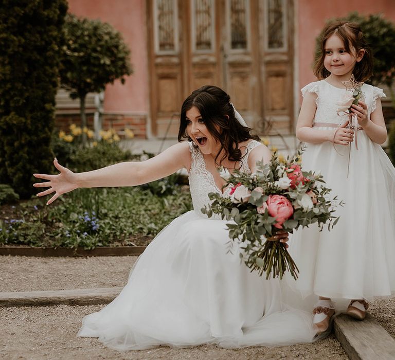 Bride in lace wedding dress and holding pink and white floral bouquet stands beside flower girl in white floor-length dress