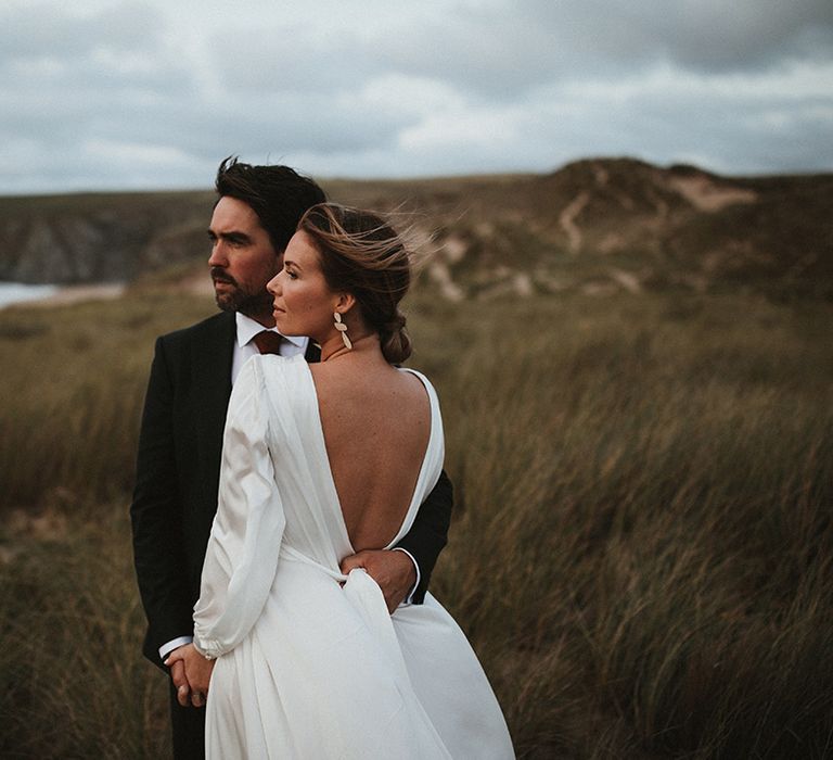 Bride in low back silk wedding dress and bell long sleeves embraces her groom outdoors beside the sea 