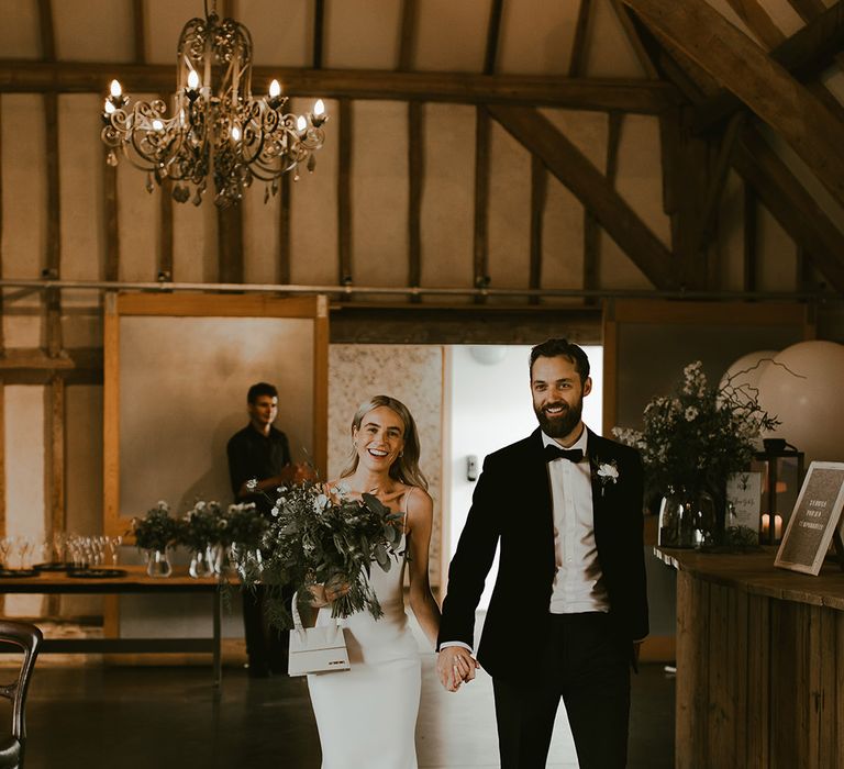 Bride in a fitted wedding dress with spaghetti straps holding a square mini Jacquemus bag and white anemone bouquet walks into the reception with the groom 