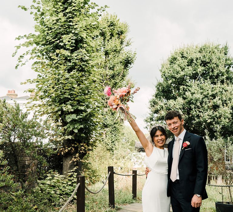 Bride in fitted silk wedding dress lifts her colourful floral bouquet in the air whilst stood beside her groom 