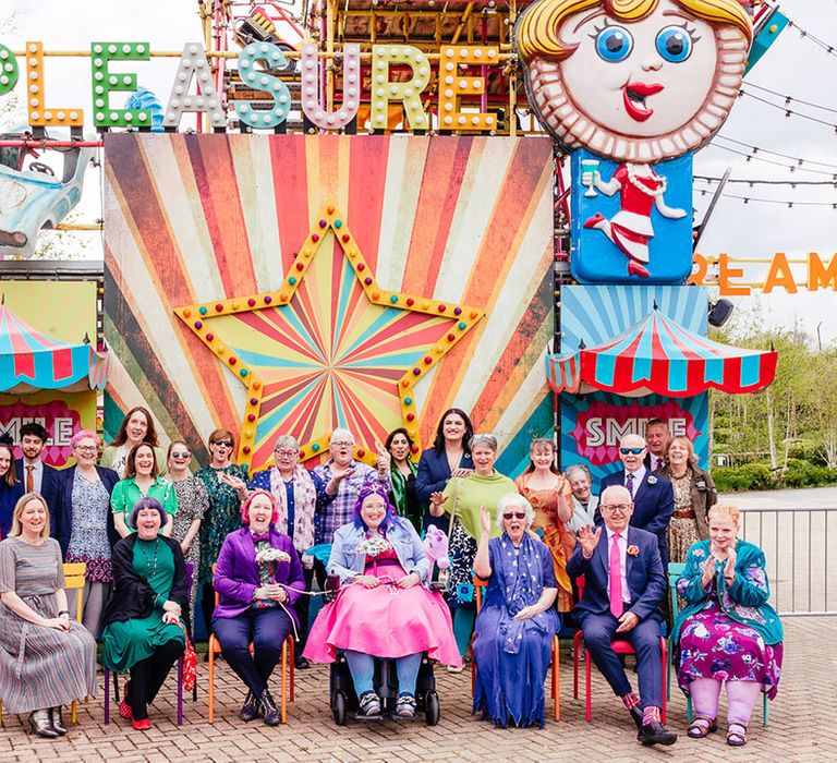 Theme park wedding with brides in complimenting pink and purple bridal outfits sitting with wedding guests outside of a large colourful rollercoaster ride at Margate Dreamland 