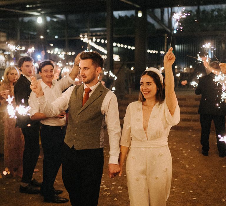 Groom in grey tweed waistcoat with an orange tie walks with the bride in a pearl flower patterned jumpsuit waving their sparklers around 