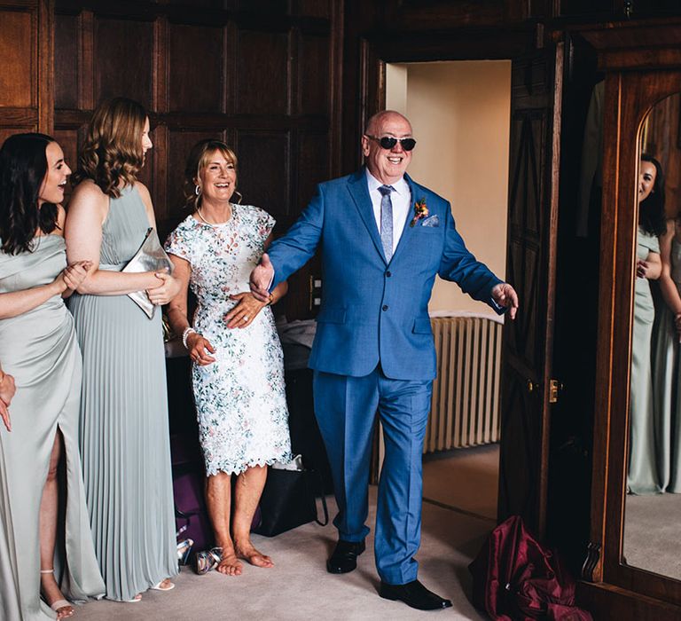 Father of the bride in a blue suit and pair of sunglasses walks into the room to get the first look at the bride 