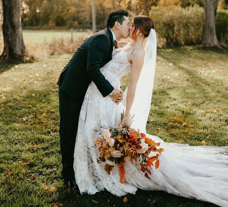 Groom dips his bride for a kiss during outdoors couples portraits as she holds orange bridal bouquet 