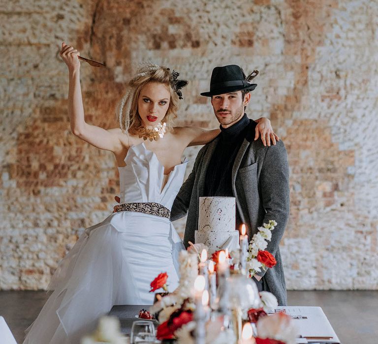 Groom in dark grey jacket, black ribbed turtle neck, black trousers and a black fedora hat and bride in organza and tulle a-line wedding day bridal ballgown with a flowing skirt, sculpted bandeau top and patterned black belt