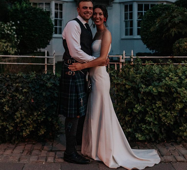 Bride in a Stephanie Allin bridal gown and the groom in a tartan kilt embrace outside their wedding venue 