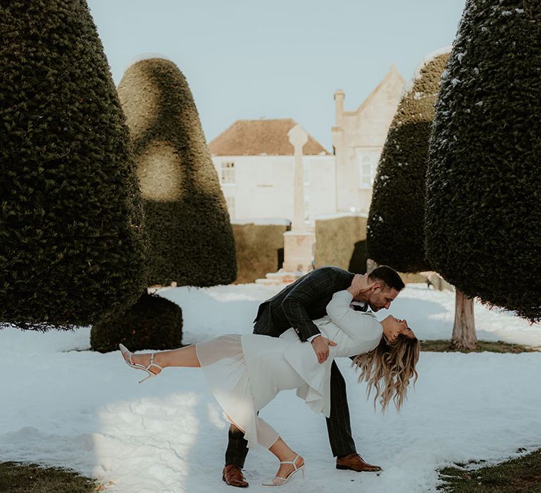 The groom in a dark grey checkered suit sweeps the bride down for a kiss in the snow 