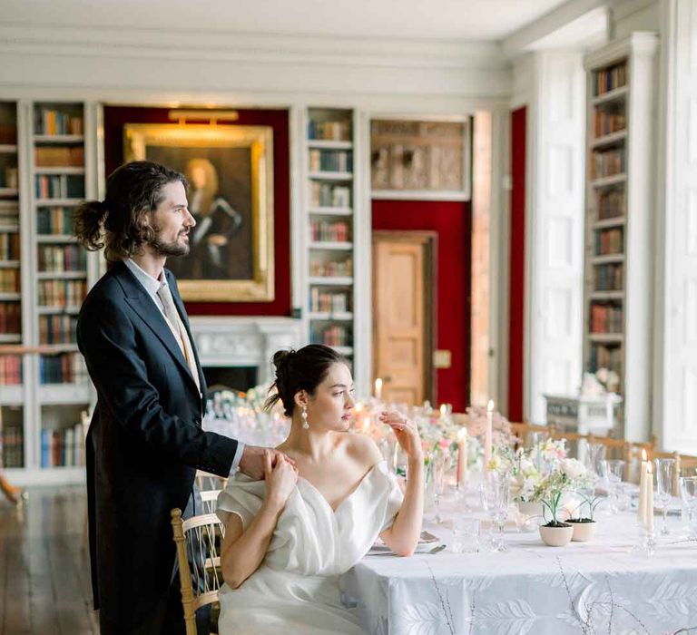 Bride wearing layered, sculpted off the shoulder wedding dress and groom in navy blue suit with yellow tie sat at classic wedding tablescape with pink tapered candles, white tablecloth and spring florals at St Giles House