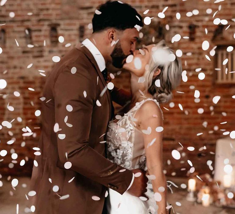 Groom in mocha brown blazer, black waistcoat, polka dot tie and pocket square and dried blush boutonniere kissing bride in  corset top sheer floral 3d appliqué wedding dress with fringe overlay with white wedding confetti falling around them 