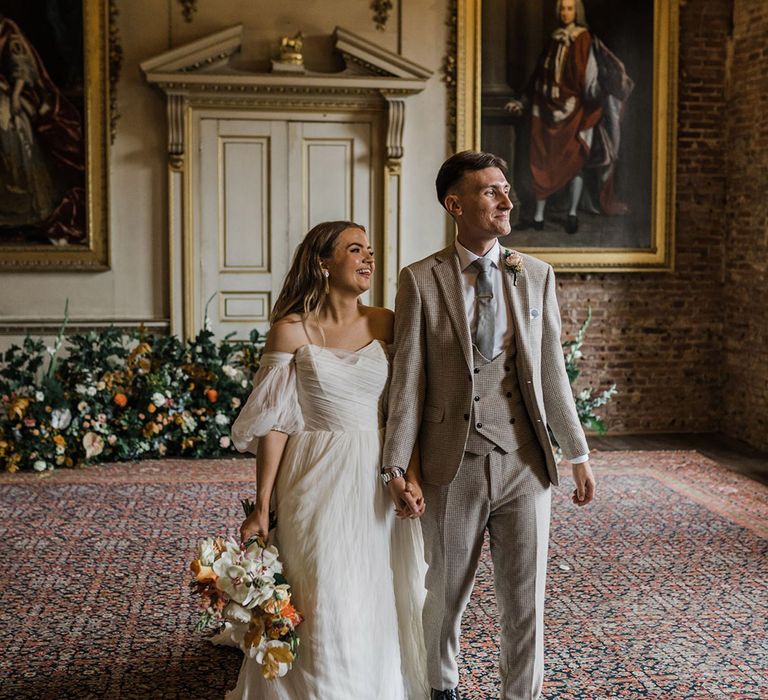 Bride in off the shoulder wedding dress with puff sleeves walking with the groom in a beige suit for their classic and elegant wedding 