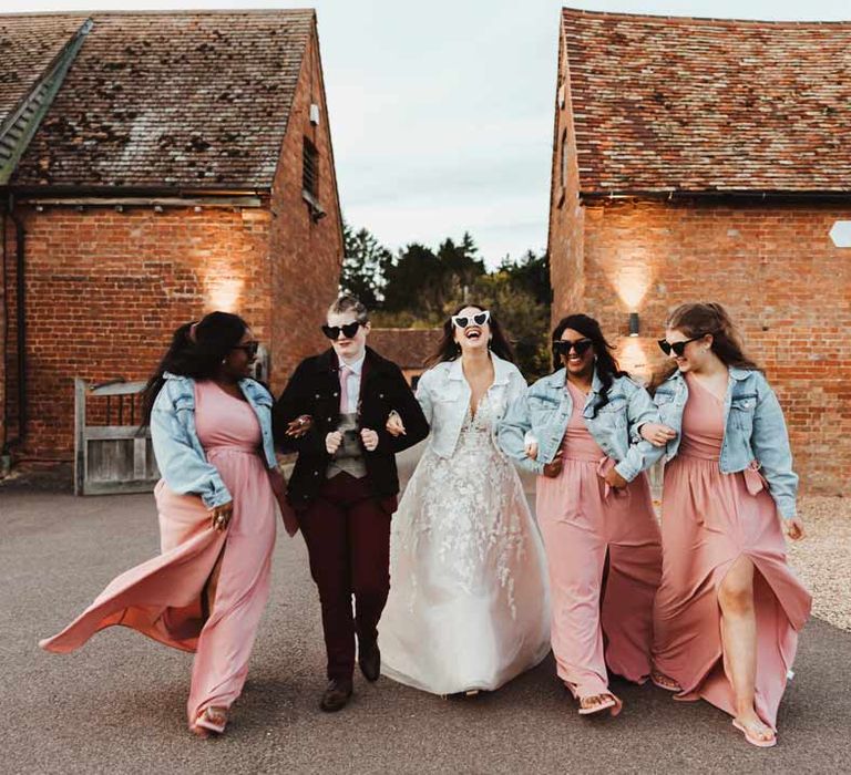 Bridal party wearing personalised denim jackets on top of dusky pink one shoulder bridesmaid dresses, illusion sleeve wedding dress and maroon bridal suit 