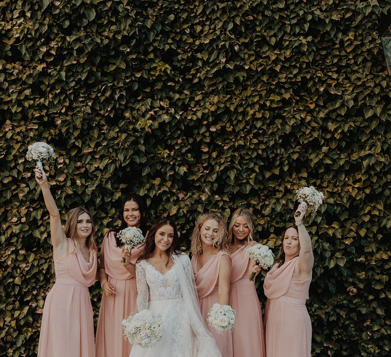 Bride in lace long sleeve Pronovias wedding dress and church-length veil holding white rose and dried flower bridal bouquet standing with bridesmaids in cowl neck short sleeve dusky pink bridesmaid dresses