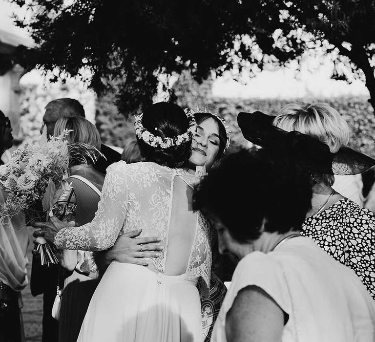 Bride in a long sleeve lace wedding dress and flower crown embracing her wedding guests 