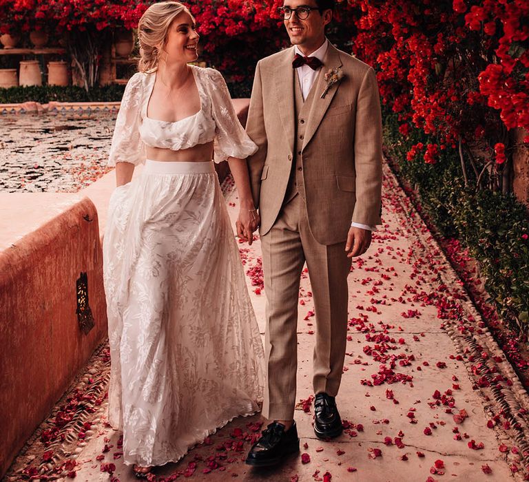 Bride in long sleeve crop top and lace maxi skirt walking hand in hand with the groom in three piece cream suit 