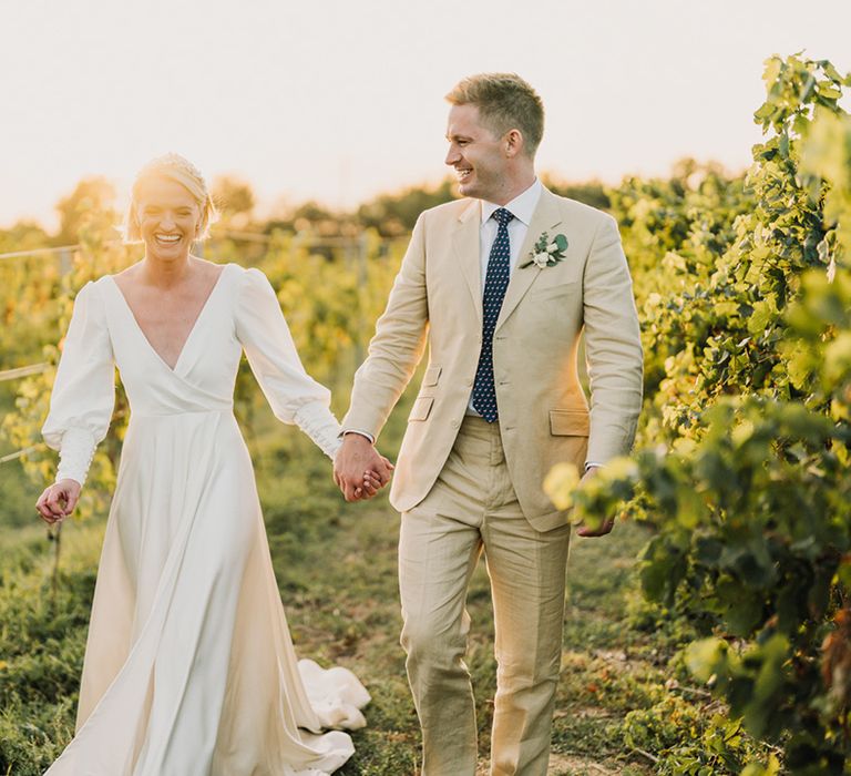 groom in a beige wedding suit holding hands with his bride in a floaty destination wedding dress